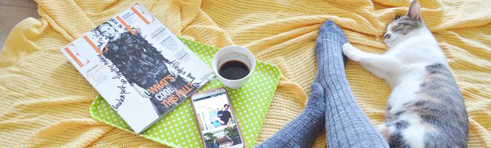 person wearing grey socks sitting on a yellow blanket with a cat, coffee, magazine and smart phone
